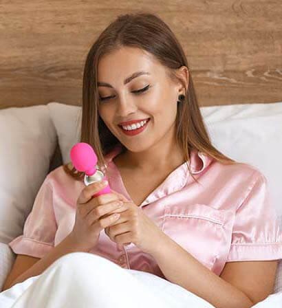 imagem mulher jovem sentada na cama com lençóis brancos segurando um vibrador varinha mágica na cor pink